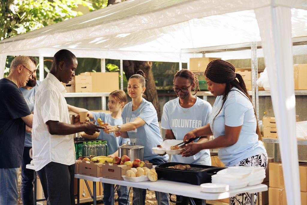 Individuals of diverse backgrounds gather outdoors to give donated food and non-perishables, providing support to the hungry and homeless. Multiracial volunteers share fresh free meals to poor people.