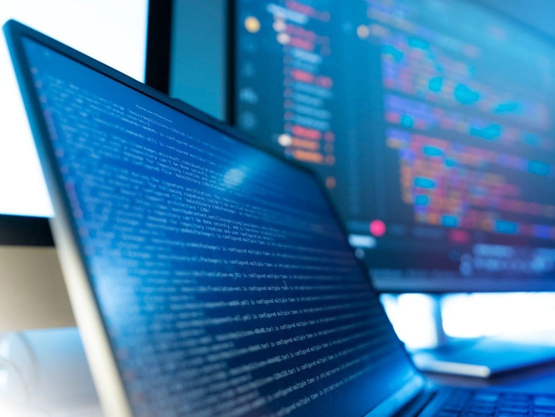 Laptop displaying blue code editor with programming text, background monitor shows blurred code, blue lighting workspace.