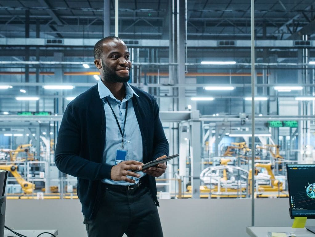 A smiling Black man in a blue shirt and dark cardigan holds a tablet in a modern factory with robotic arms.