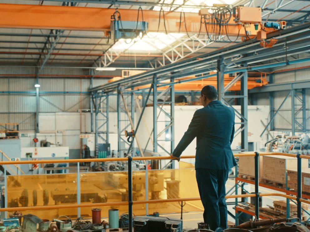 A man in a blue suit leans on a railing in a large factory, viewing machinery and an orange overhead crane.