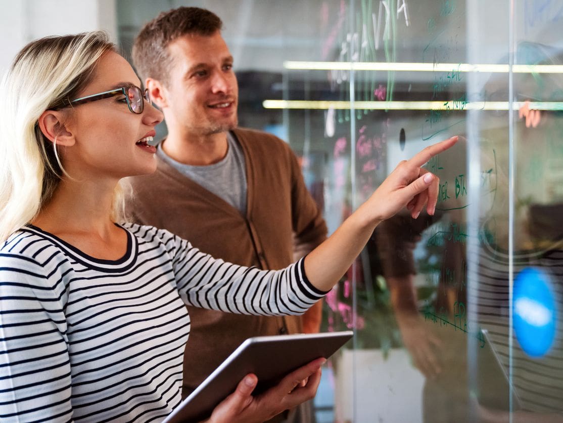 Blonde woman in striped shirt and glasses points at glass whiteboard while holding a tablet, man smiles behind her in office.