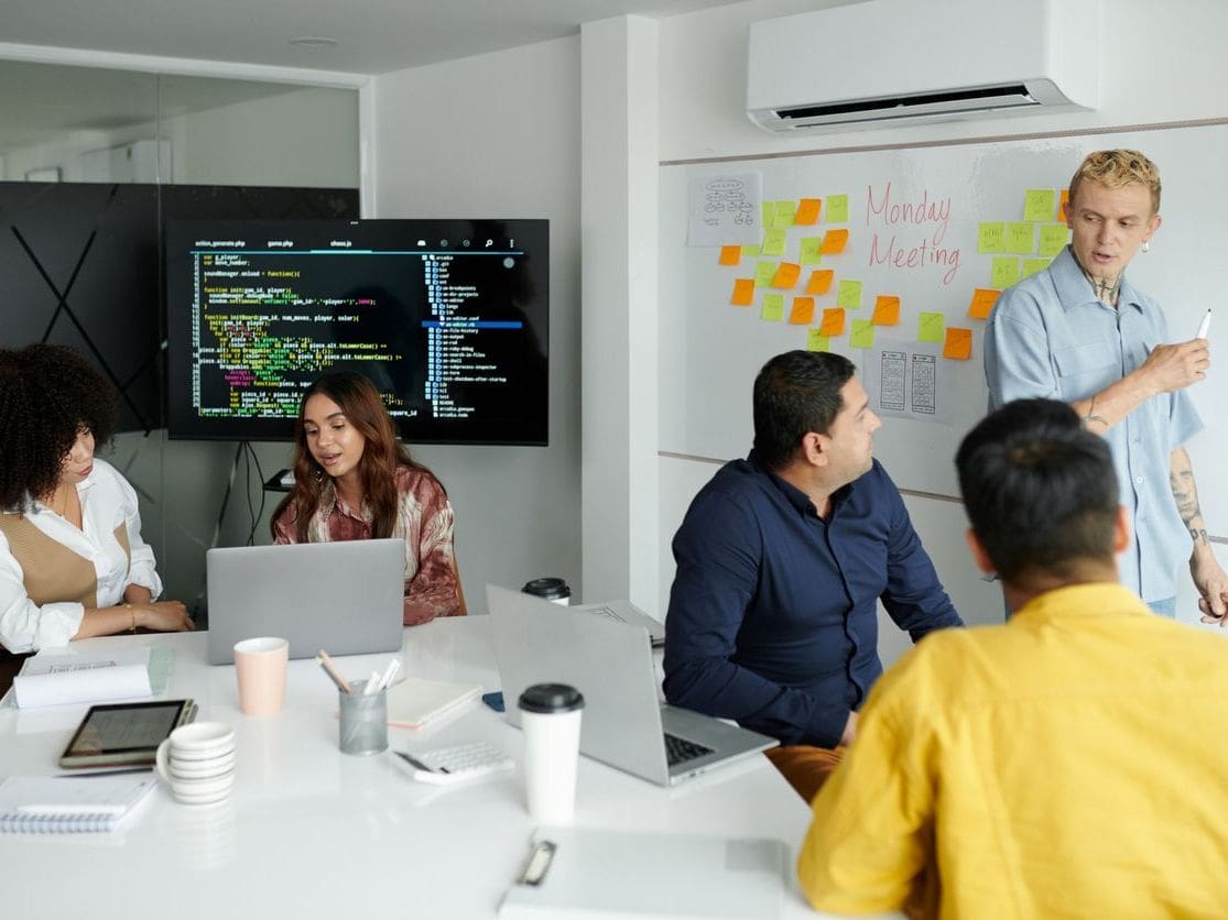 Team meeting in modern office, developers reviewing code on screen, presenter writing on whiteboard with sticky notes.