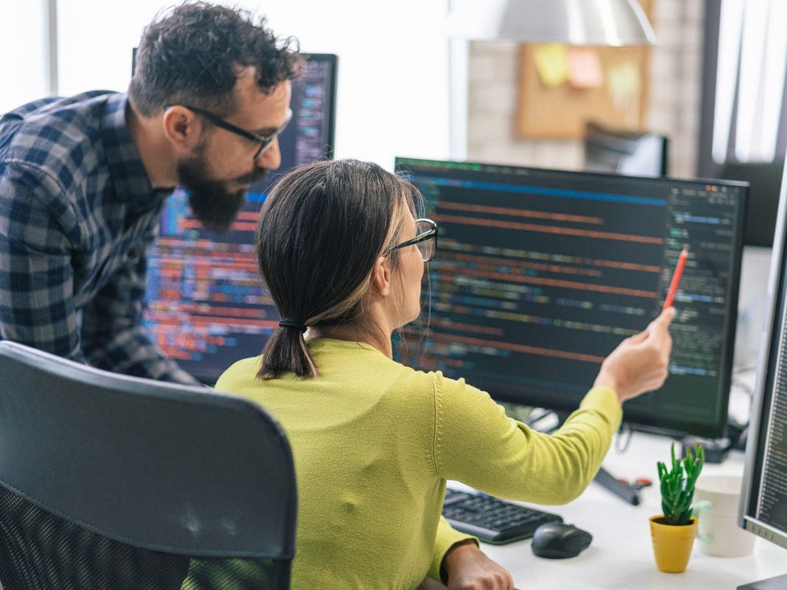 Two developers work together; woman in yellow points at code on monitor with red pen while man in plaid looks on.