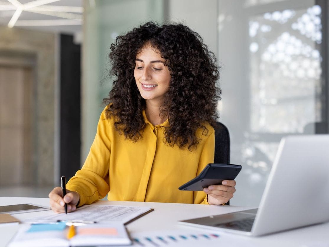 Curly‑haired woman in yellow shirt writes on paper with pen, holds tablet, laptop on desk in office.